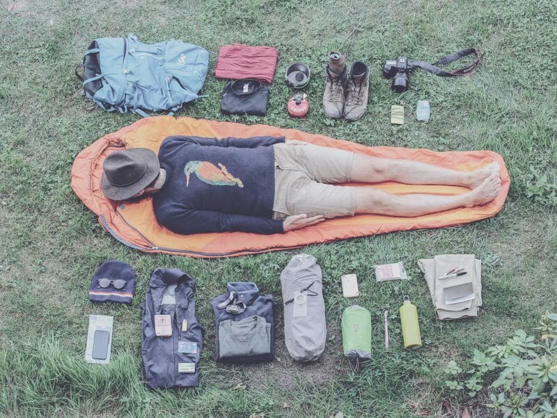 un homme allongé sur un sac de couchage dans l'herbe entouré d'équipements de rando